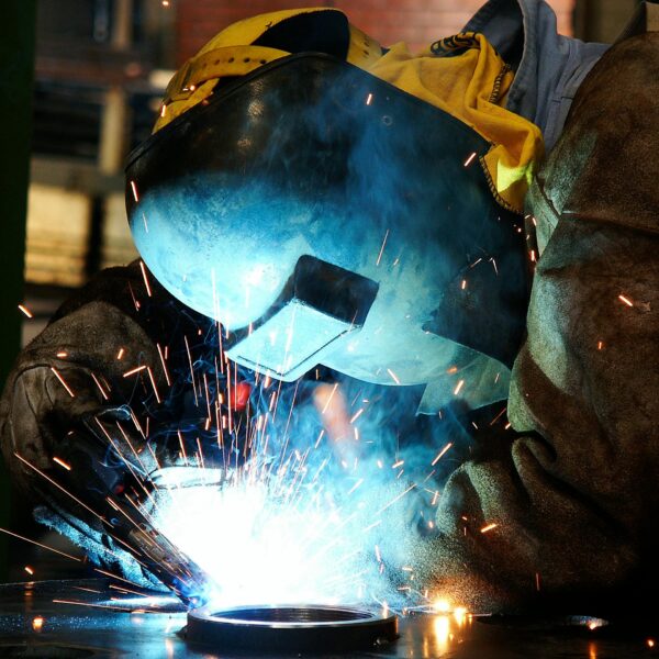 A welder in protective gear creates sparks while working on metal indoors.